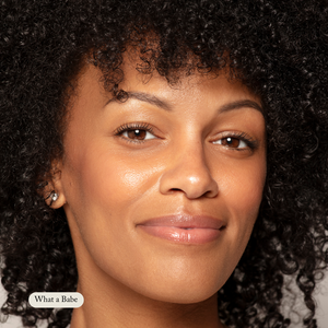 A woman with curly hair and clear skin smiles softly at the camera against a plain background. In the corner, a text box says “What a Babe,” highlighting her natural glow enhanced by Toups and Co Organics’ Organic Lip Gloss.