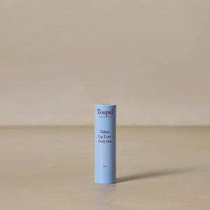 A single tube of Toups and Co Organics Candy Cane Lip Love, crafted with nourishing grass-fed tallow, stands upright on a beige surface against a neutral background.