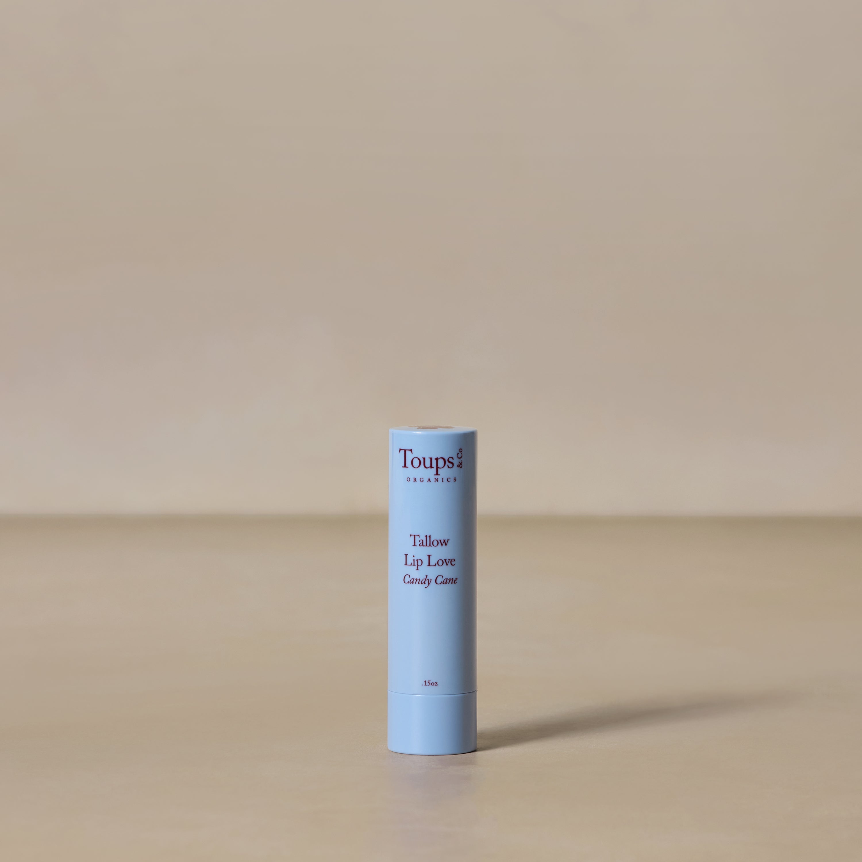 A tube of Toups and Co Organics Candy Cane Lip Love stands upright on a neutral beige surface with a matching background, showcasing its nourishing benefits.