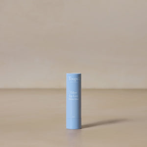 A light blue tube of Toups and Co Organics Tallow Lip Love hydrating lip balm stands upright on a beige surface against a neutral background.