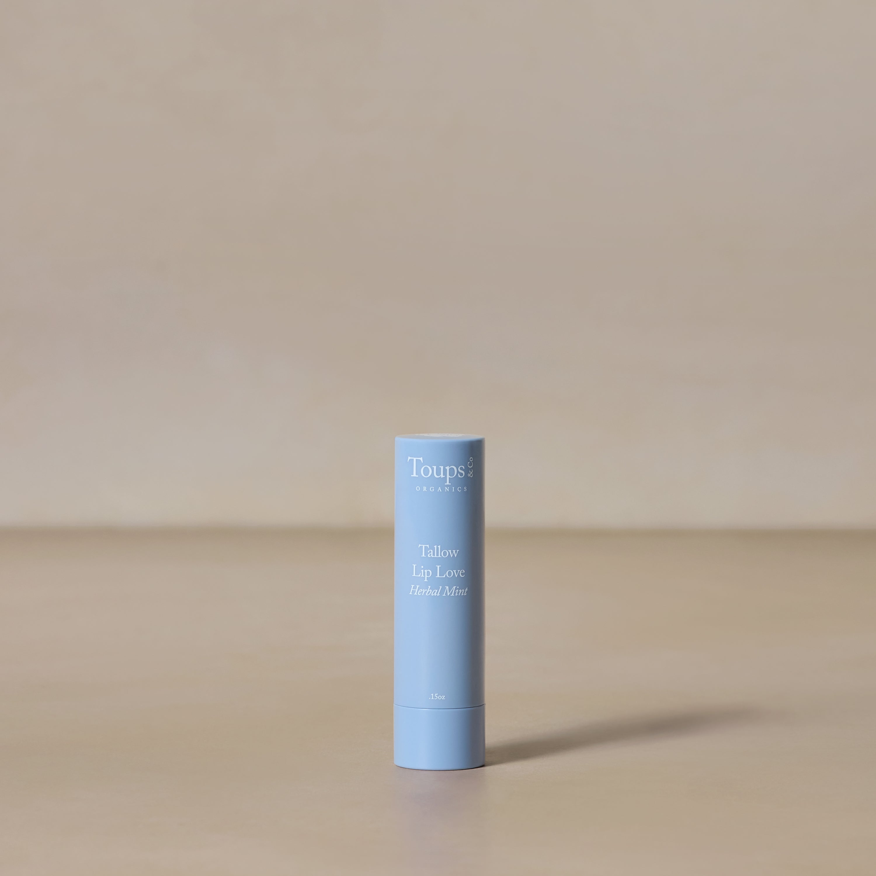 A light blue tube of Toups and Co Organics Tallow Lip Love stands upright on a neutral beige surface with a plain beige background, ideal for soothing chapped lips.