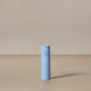 A light blue tube of Toups and Co Organics Tallow Lip Love hydrating lip balm stands upright on a beige surface with a neutral background.