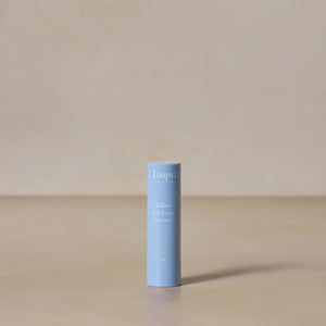 A blue tube of Toups and Co Organics Tallow Lip Love Unscented hydrating lip balm stands upright on a neutral beige surface with a plain beige background.