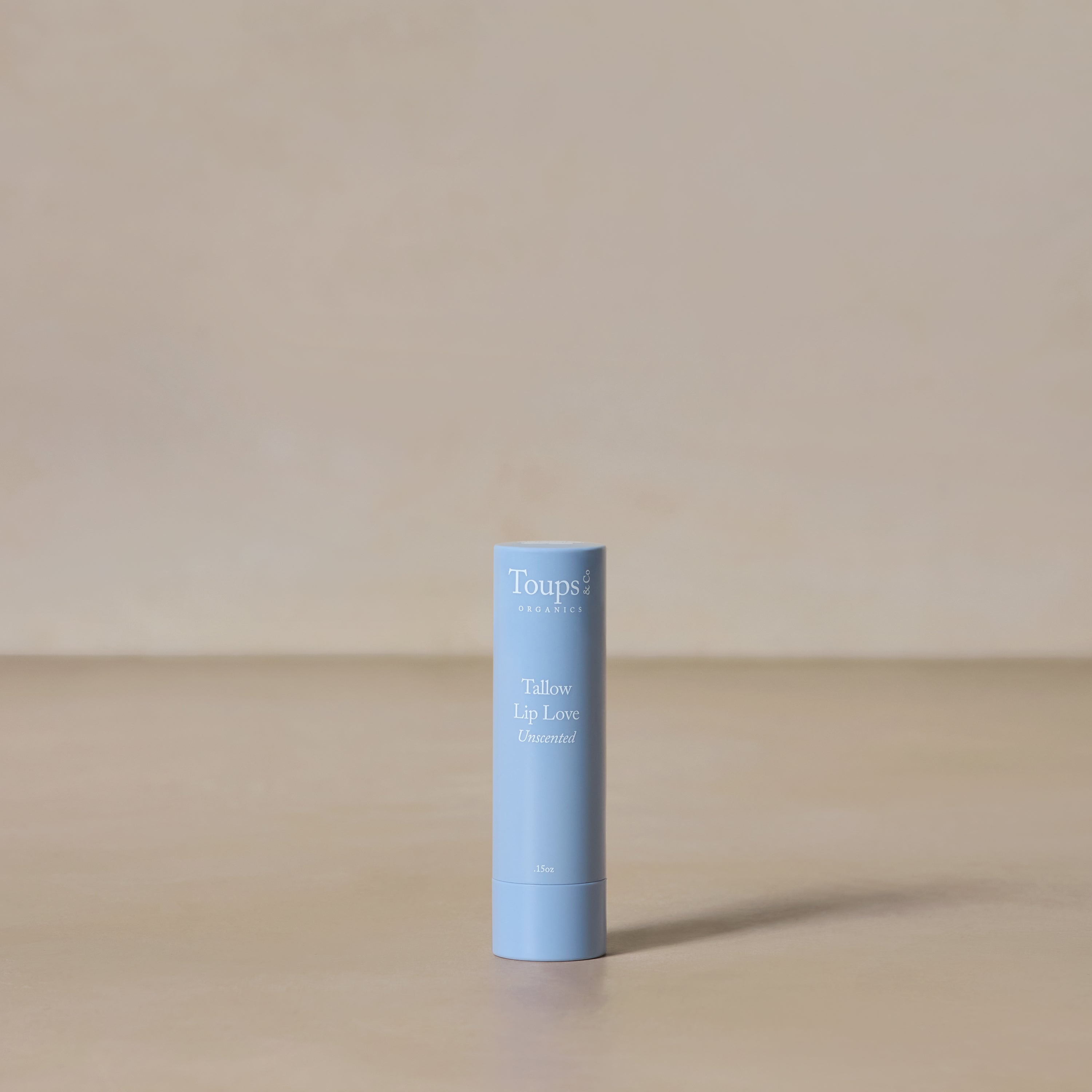 A blue tube of Toups and Co Organics Tallow Lip Love Unscented hydrating lip balm stands upright on a neutral beige surface with a plain beige background.