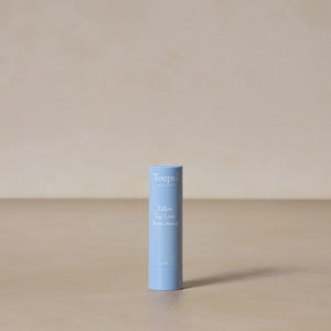 A blue tube of Toups and Co Organics Tallow Lip Love stands upright on a neutral beige surface with a plain beige background, ideal for soothing chapped lips.