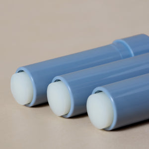 Three blue cylindrical tubes of Tallow Lip Love by Toups and Co Organics, each with white rounded caps, are arranged parallel on a beige surface.