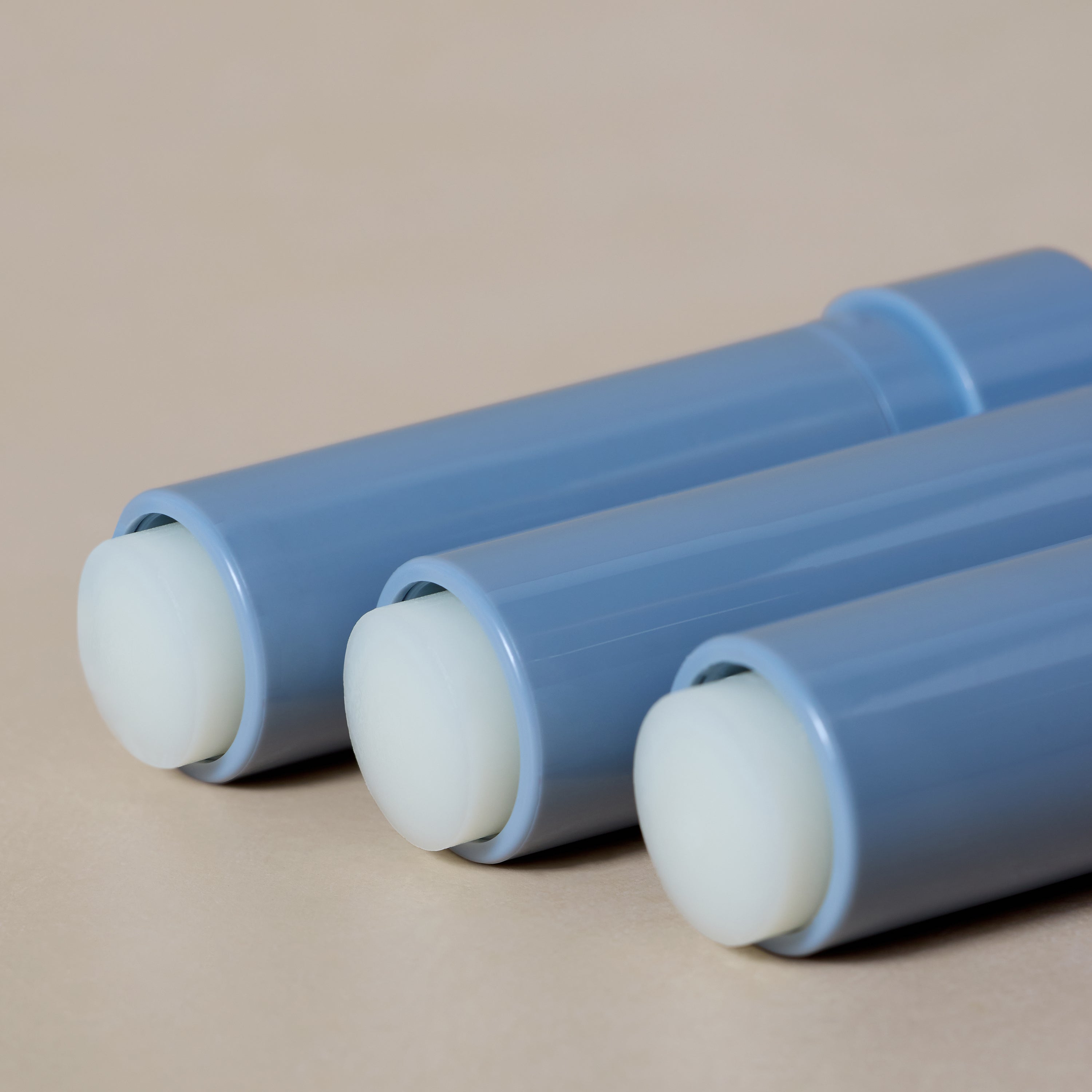 Three blue cylindrical tubes of Tallow Lip Love by Toups and Co Organics, each with white rounded caps, are arranged parallel on a beige surface.