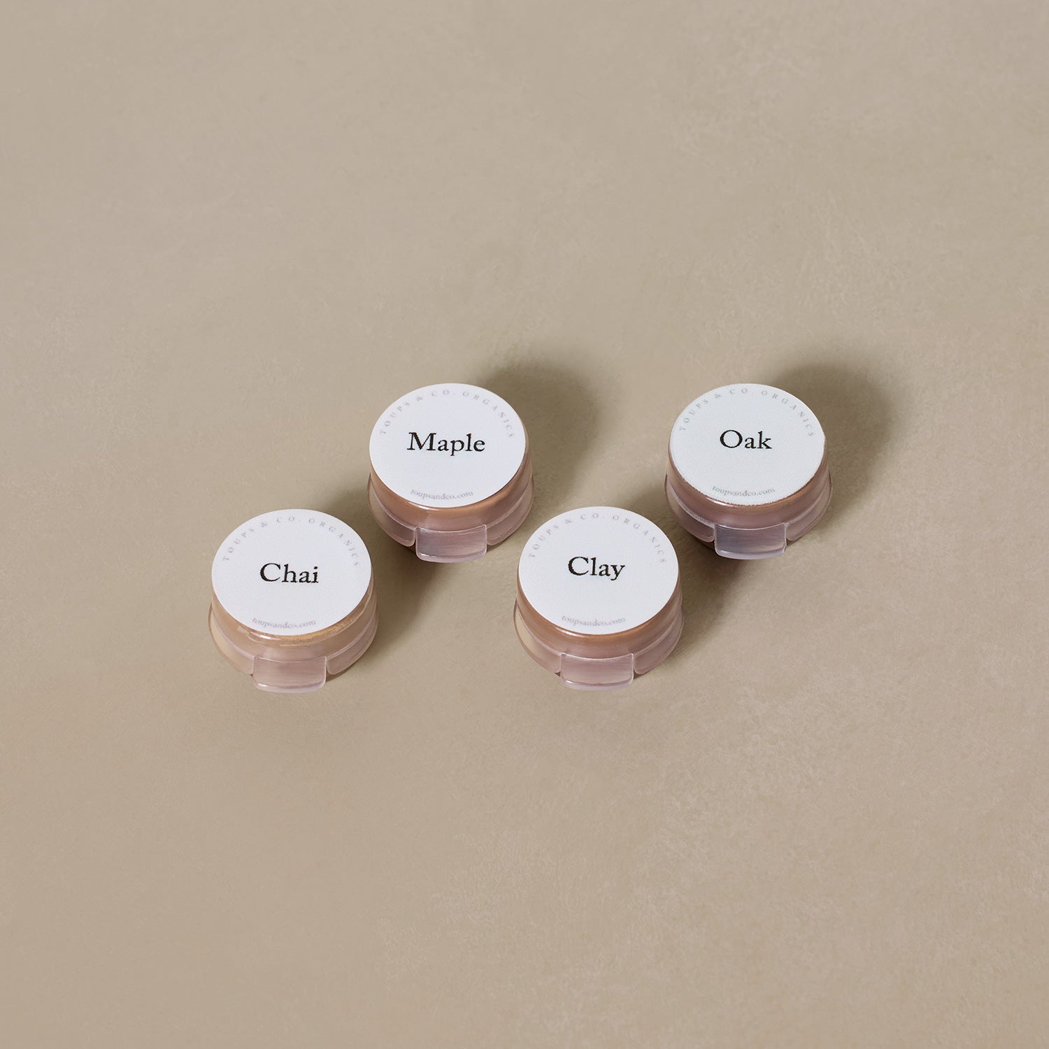 Four small containers labeled Chai, Maple, Oak, and Clay from the Toups and Co Organics Sample Set - Liquid Foundation are arranged on a neutral beige surface, highlighting our all-natural formula.