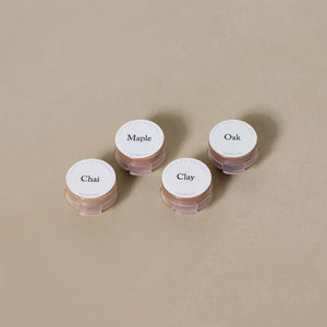 Four small containers labeled Chai, Maple, Oak, and Clay from the Toups and Co Organics Sample Set - Liquid Foundation are arranged on a neutral beige surface, highlighting our all-natural formula.
