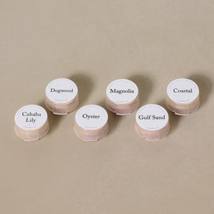 Six small round containers from the Toups and Co Organics Sample Set - Liquid Foundation, labeled Dogwood, Magnolia, Coastal, Cahaba Lily, Oyster, and Gulf Sand, are arranged on a beige surface—ideal for sampling foundation shades.