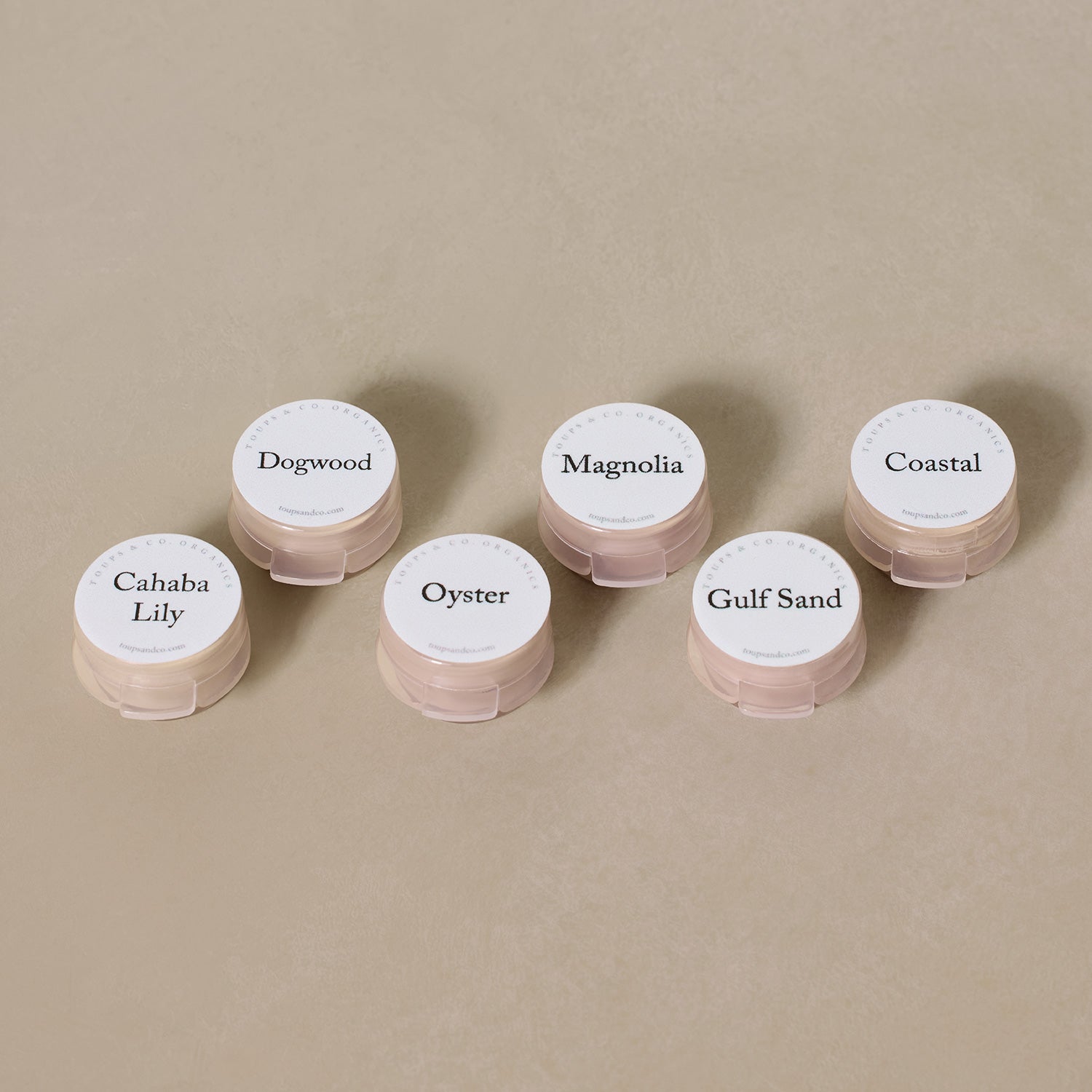 Six small containers labeled Dogwood, Magnolia, Coastal, Cahaba Lily, Oyster, and Gulf Sand—a Sample Set - Liquid Foundation from Toups and Co Organics—are arranged on a beige surface.