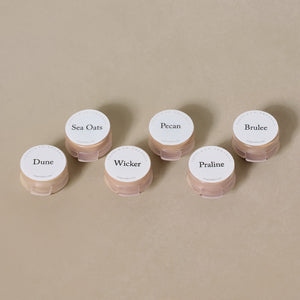 Six round containers labeled Dune, Sea Oats, Wicker, Pecan, Praline, and Brulee rest on a neutral surface—the Toups and Co Organics Sample Set - Liquid Foundation made with all-natural ingredients.