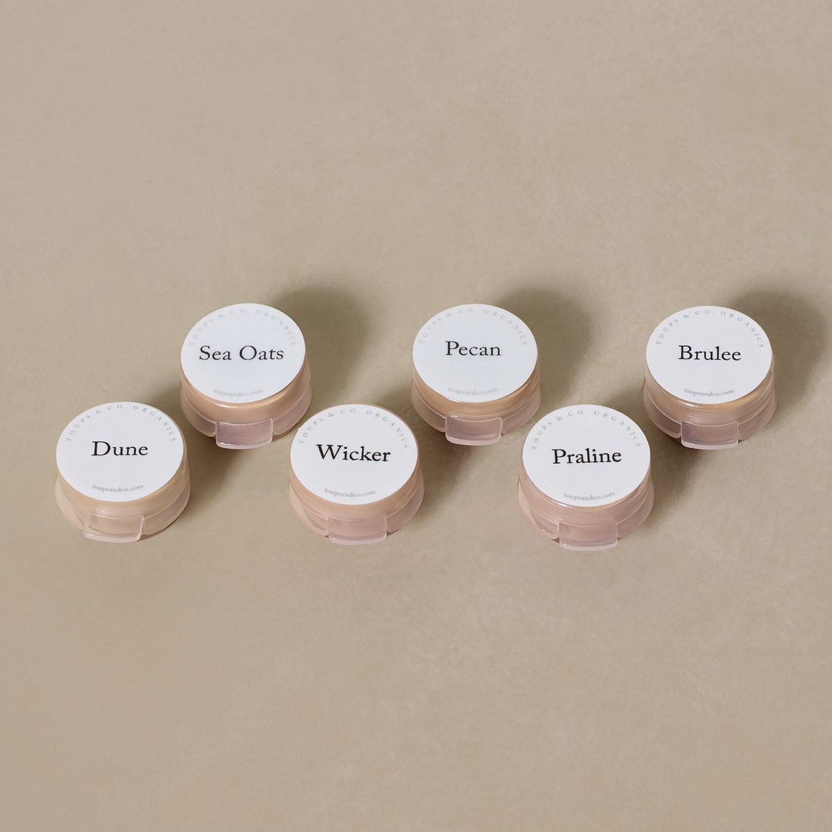 Six round containers labeled Dune, Sea Oats, Wicker, Pecan, Praline, and Brulee rest on a neutral surface—the Toups and Co Organics Sample Set - Liquid Foundation made with all-natural ingredients.