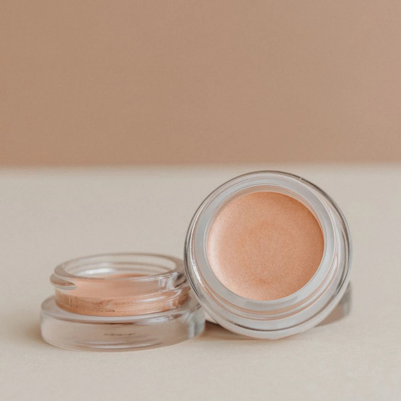 Two small glass pots of shimmering peach-toned cream makeup.