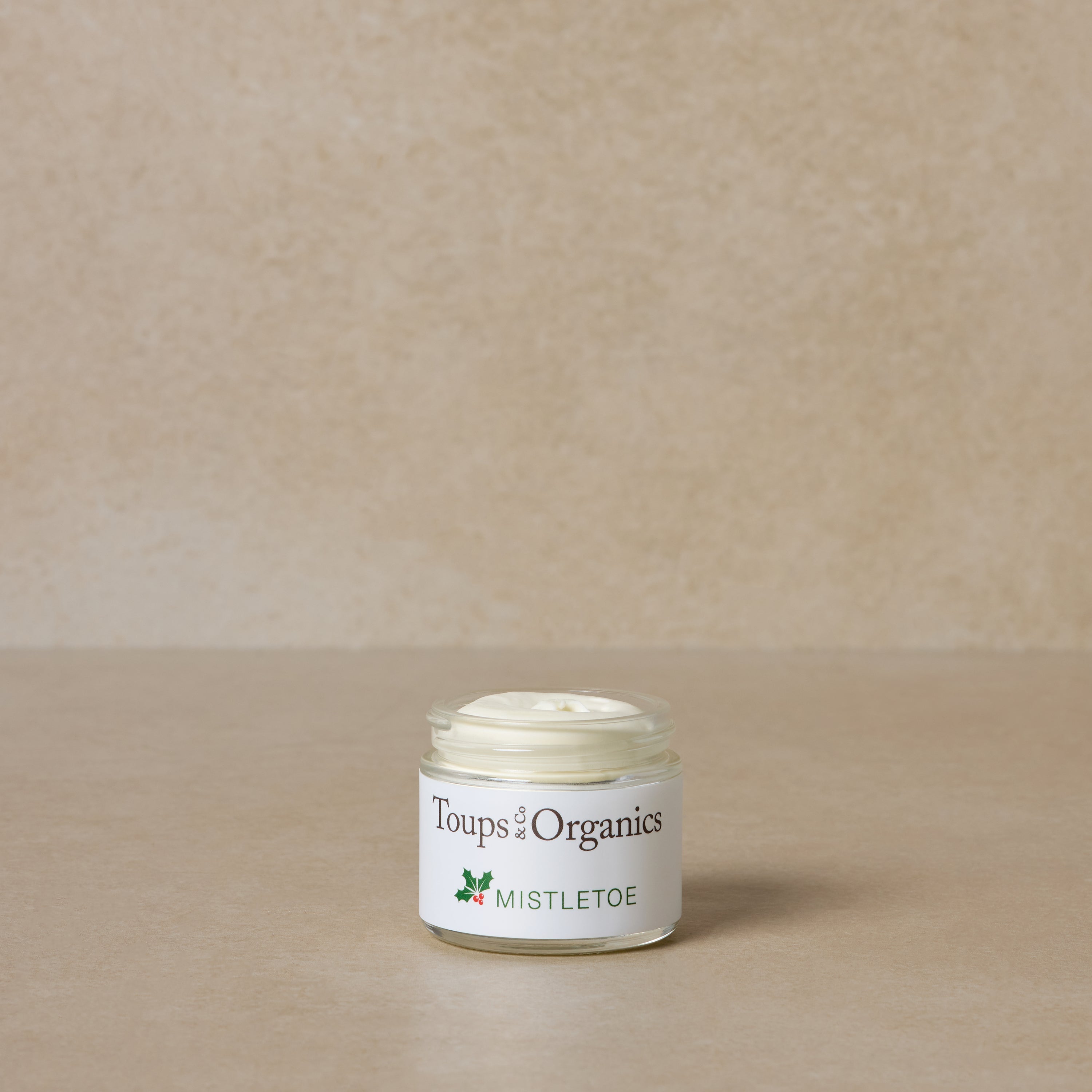 A glass jar labeled Toups and Co Organics Mistletoe Balm sits on a beige surface with a matching background—made with grass-fed tallow, it's perfect for thoughtful Christmas gifts.