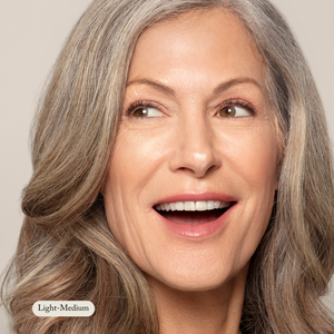 An older woman with long gray hair, clear skin, and a joyful expression looks upward. A Light-Medium label appears in the corner. She wears Toups and Co Organics Mineral Makeup with natural SPF for healthy, glowing skin.