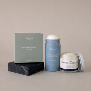 Three products from Toups and Co Organics’ Non Toxic Starter Set—a box labeled Activated Charcoal Face Bar, a stick of Natural Deodorant, and an open jar of Unscented Tallow Balm—are displayed on a neutral background.