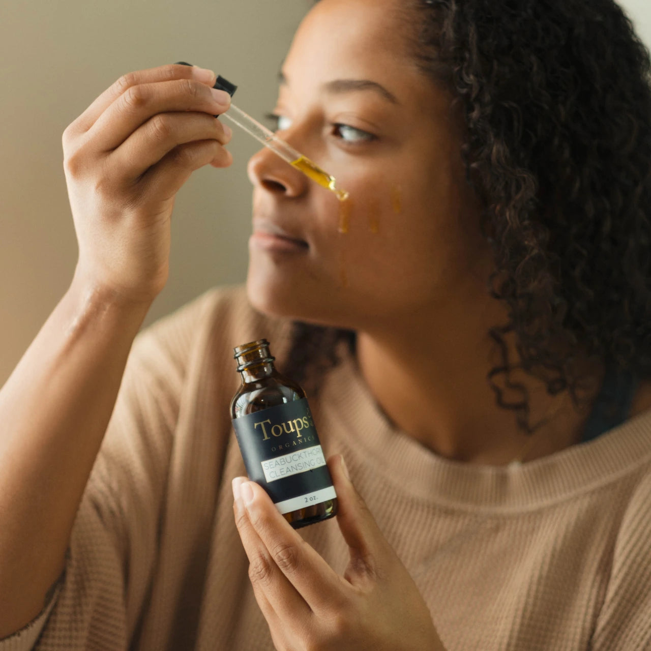A person applying Toups Organics Seabuckthorn Cleansing Oil from a dropper bottle onto their cheek.