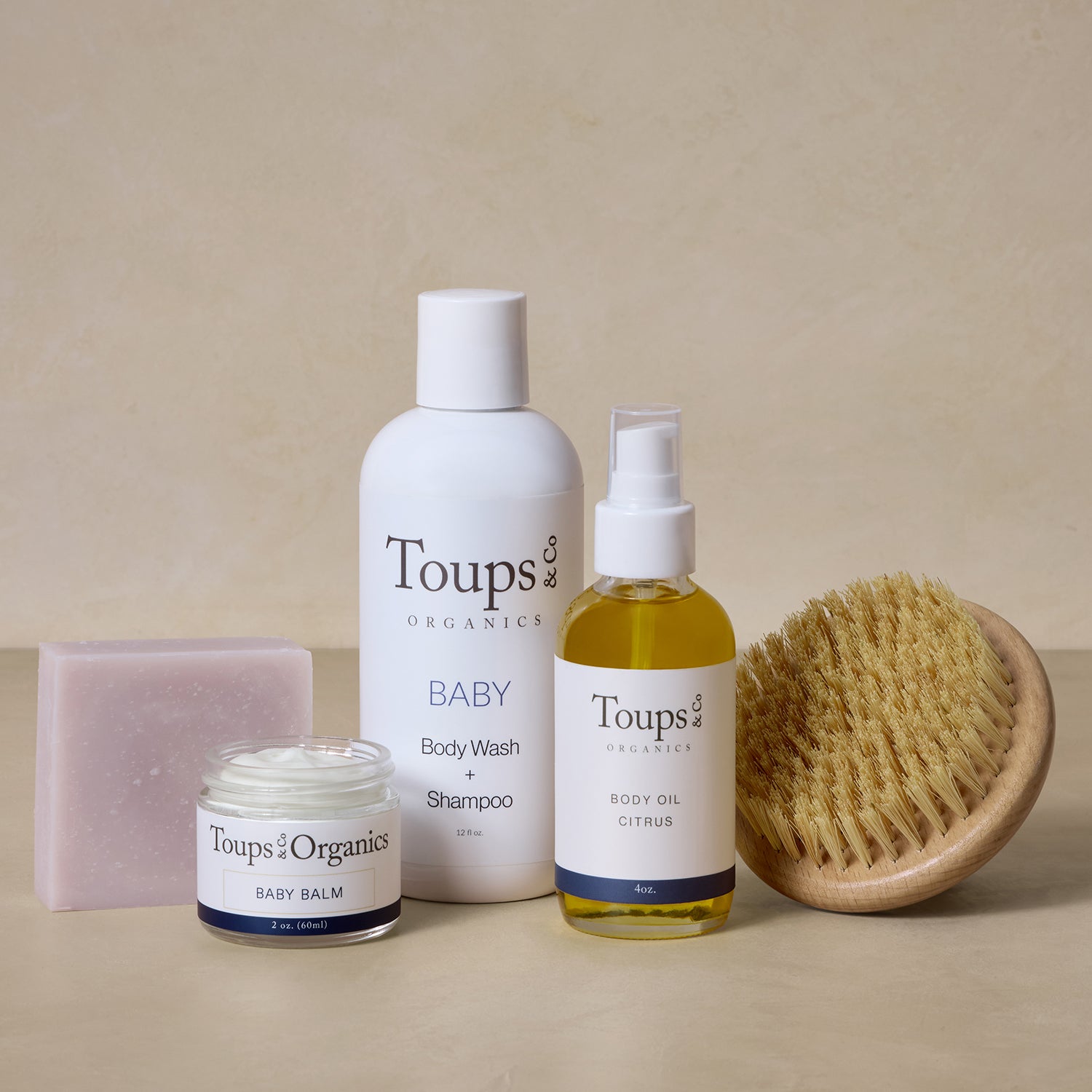The Postpartum Skincare Set by Toups and Co Organics, featuring soap, baby balm, body wash, body oil, and a dry brush, is arranged on a beige surface.