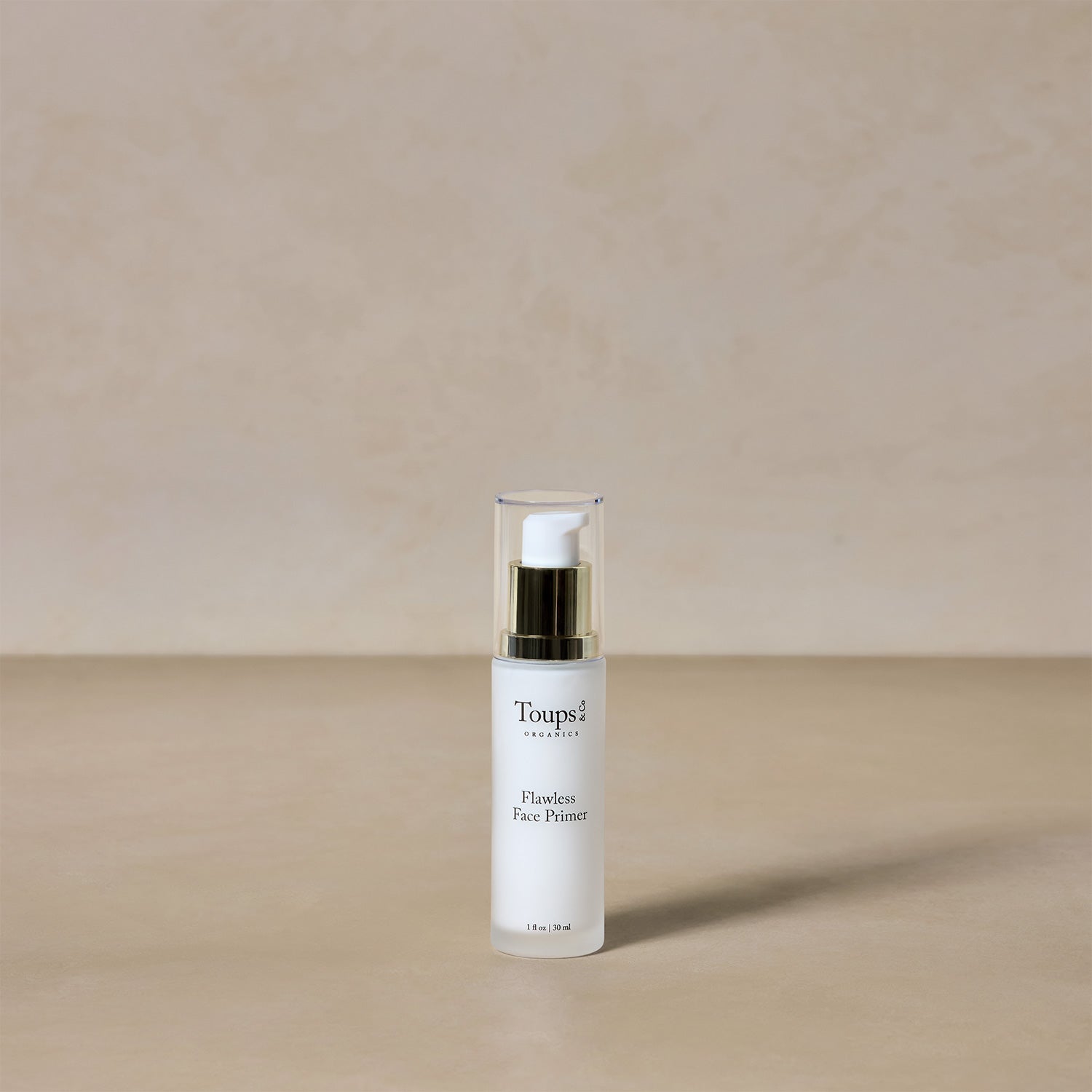 A bottle of Toups and Co Organics Flawless Face Primer, an antioxidant-rich primer with a pump dispenser, rests on a beige surface against a light beige background.
