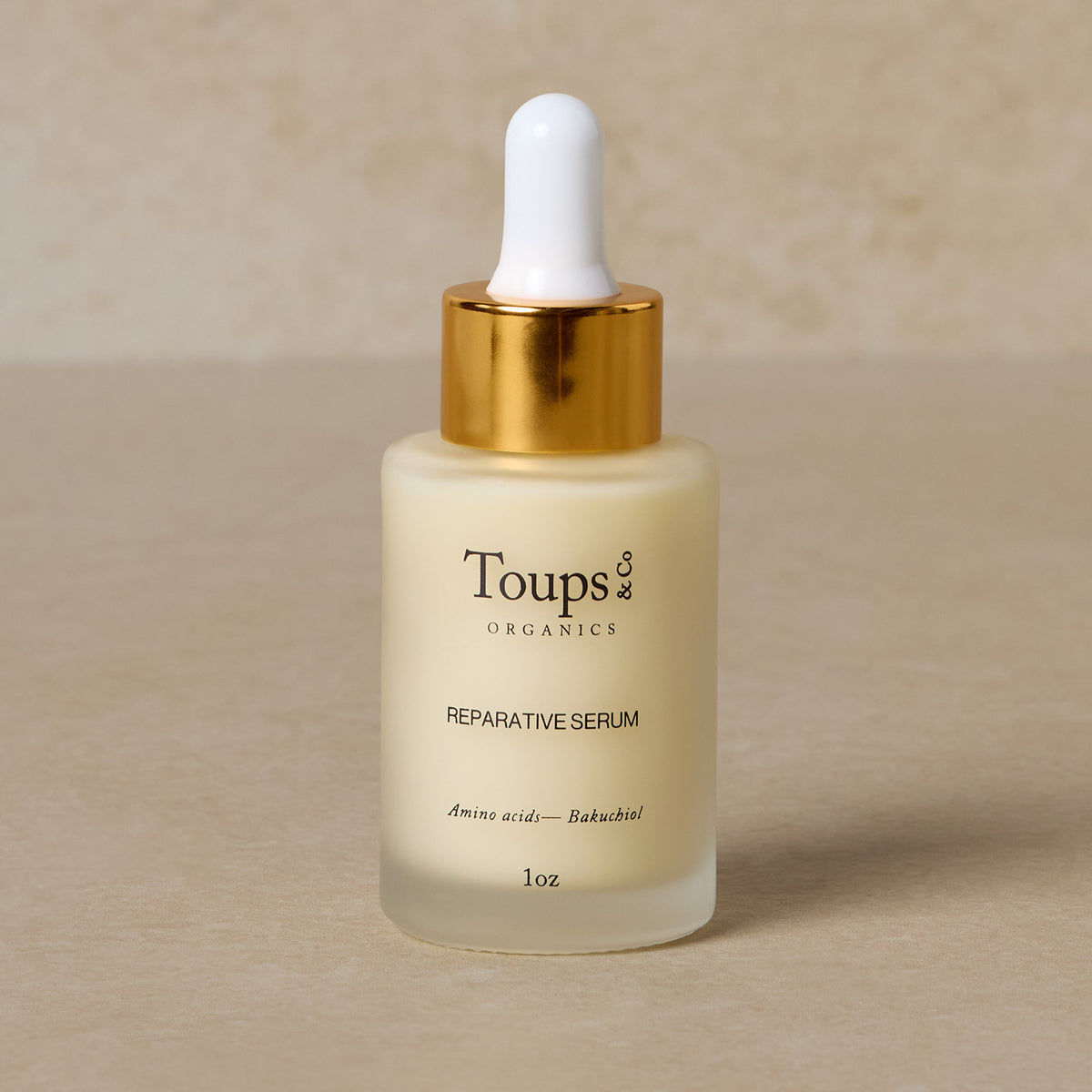 A frosted glass dropper bottle with a gold cap, labeled Toups and Co Organics Reparative Serum, ideal for adding a bakuchiol serum to your anti-aging routine, elegantly sits on a neutral surface.