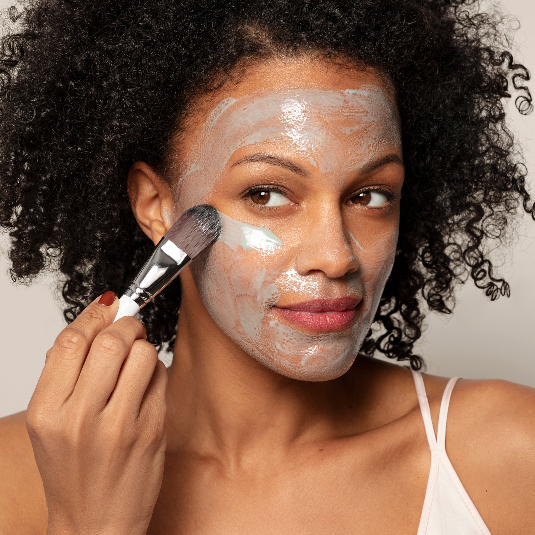 A woman in a white camisole applies Toups and Co Organics Resurfacing Mask with blue C-phycocyanin algae extract to her cheek using a brush, looking to the side.