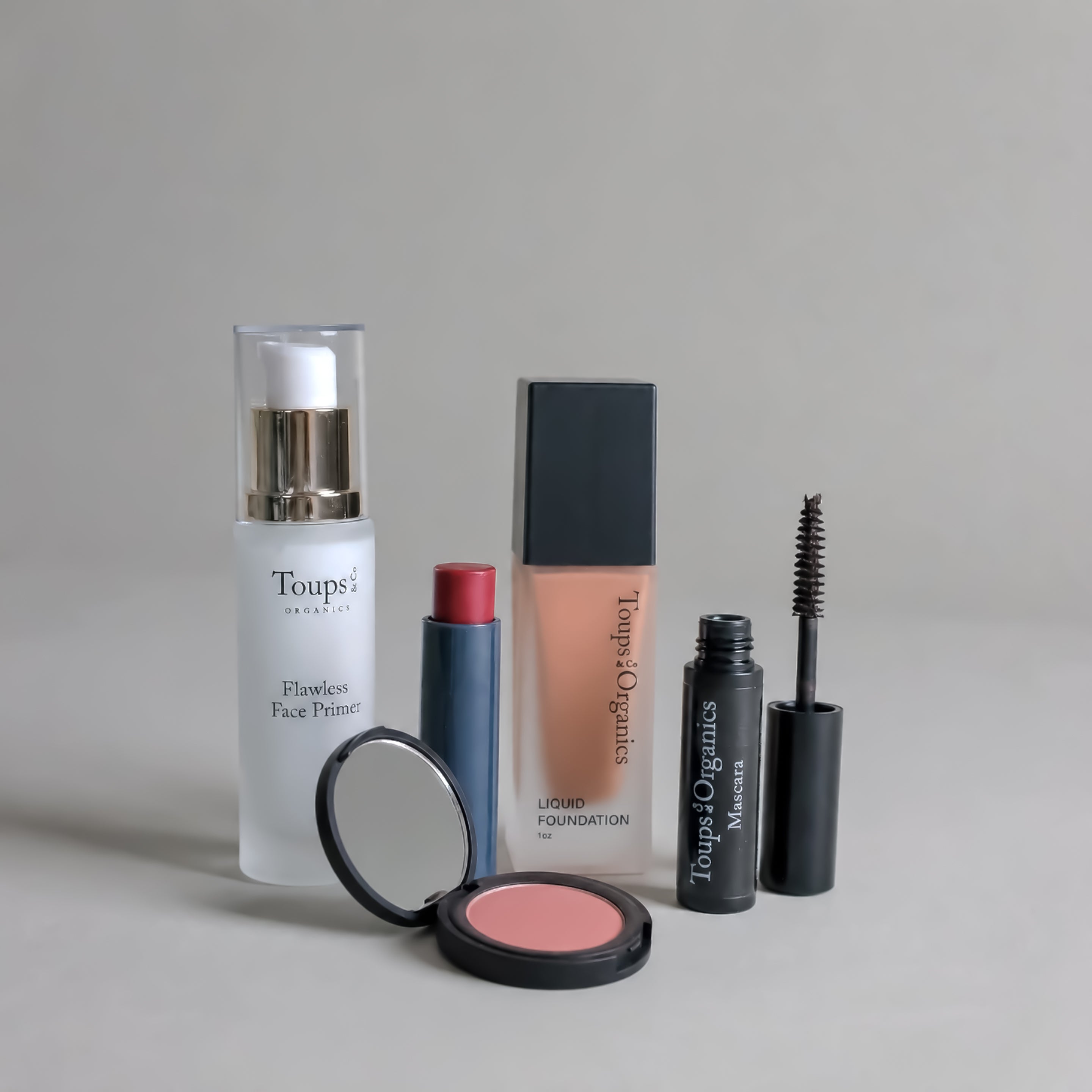 Five products from the Toups and Co Organics Complete Makeup Set—face primer, liquid foundation, blush tinted lip balm, and mascara with an open wand—are arranged on a flat surface.