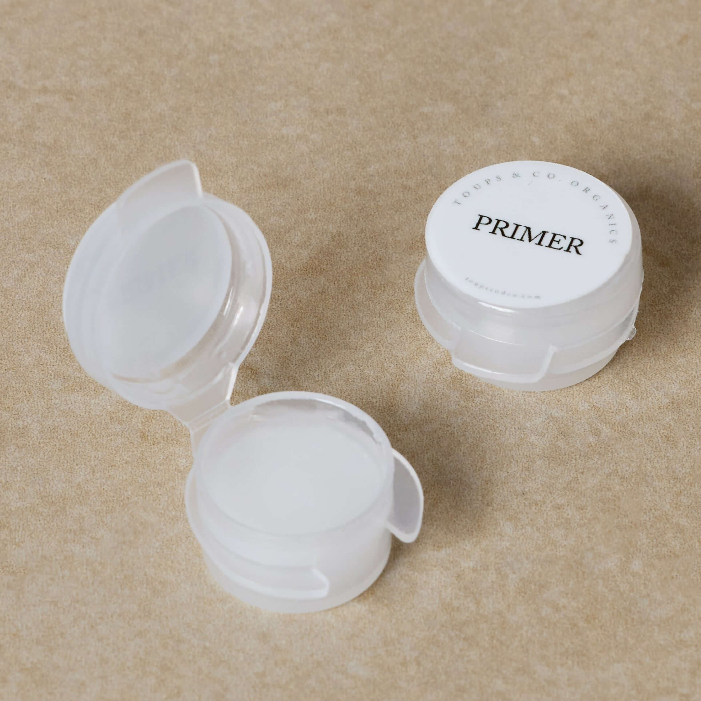 Two small round containers of Toups and Co Organics Sample - Flawless Face Primer are displayed, one open and one closed, on a tan textured surface. The closed container is labeled PRIMER for smooth makeup application.