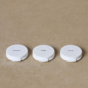 Three round white containers from the Toups and Co Organics Sample Set - Mineral Foundation, labeled Caramel, Toffee, and Cocoa, are arranged in a row on a beige surface.