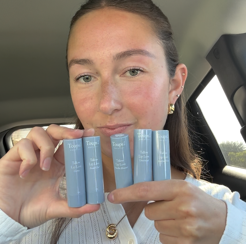 A person in a car holds four tubes of Toups and Co Organics Tallow Lip Love hydrating lip balm, made with grass-fed tallow, displaying the labels towards the camera.