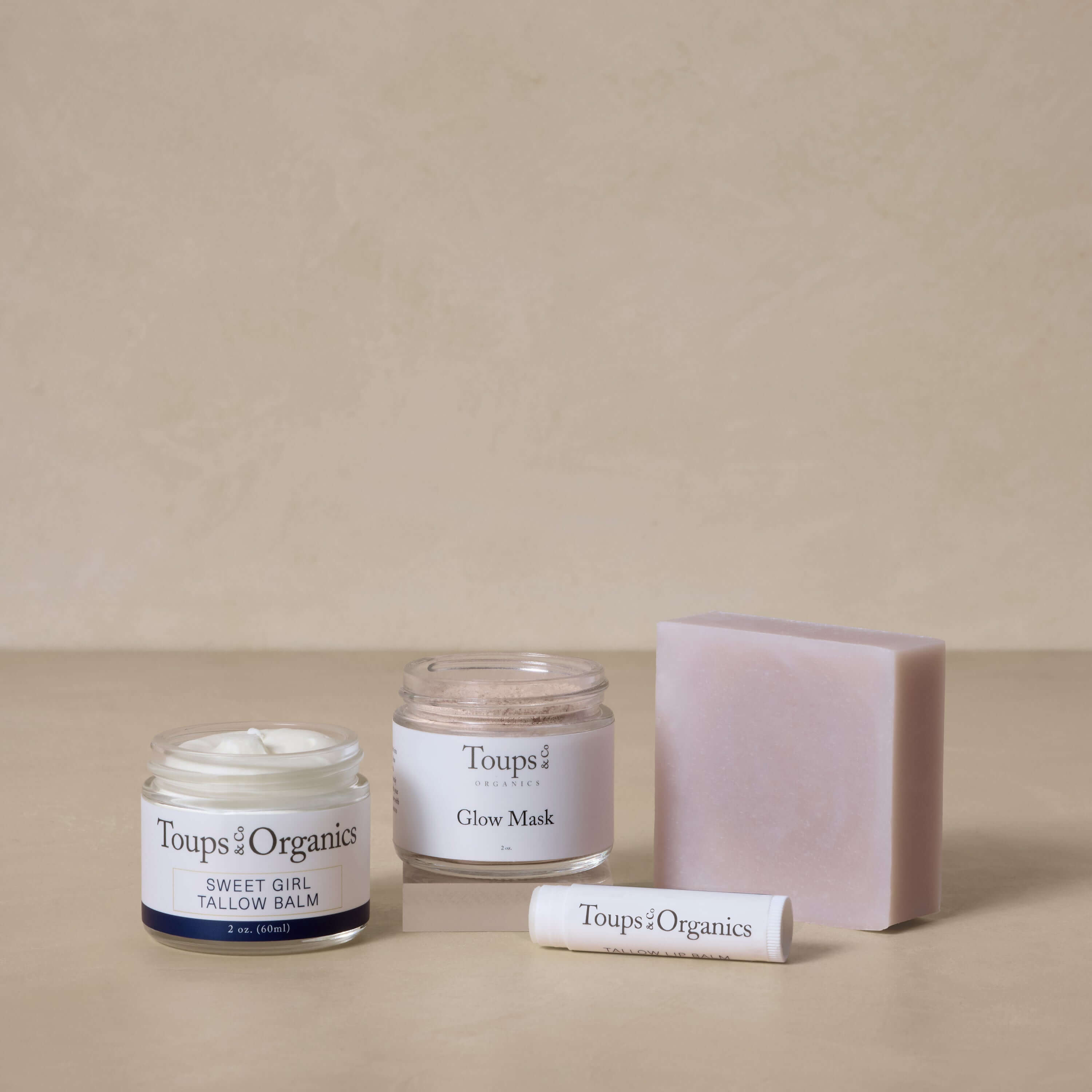 The Toups and Co Organics Spa Gift Set, featuring Sweet Girl Tallow Balm, Glow Mask, lip balm, and soap, is arranged on a neutral surface with a beige background—an ideal gift for nontoxic skincare lovers.