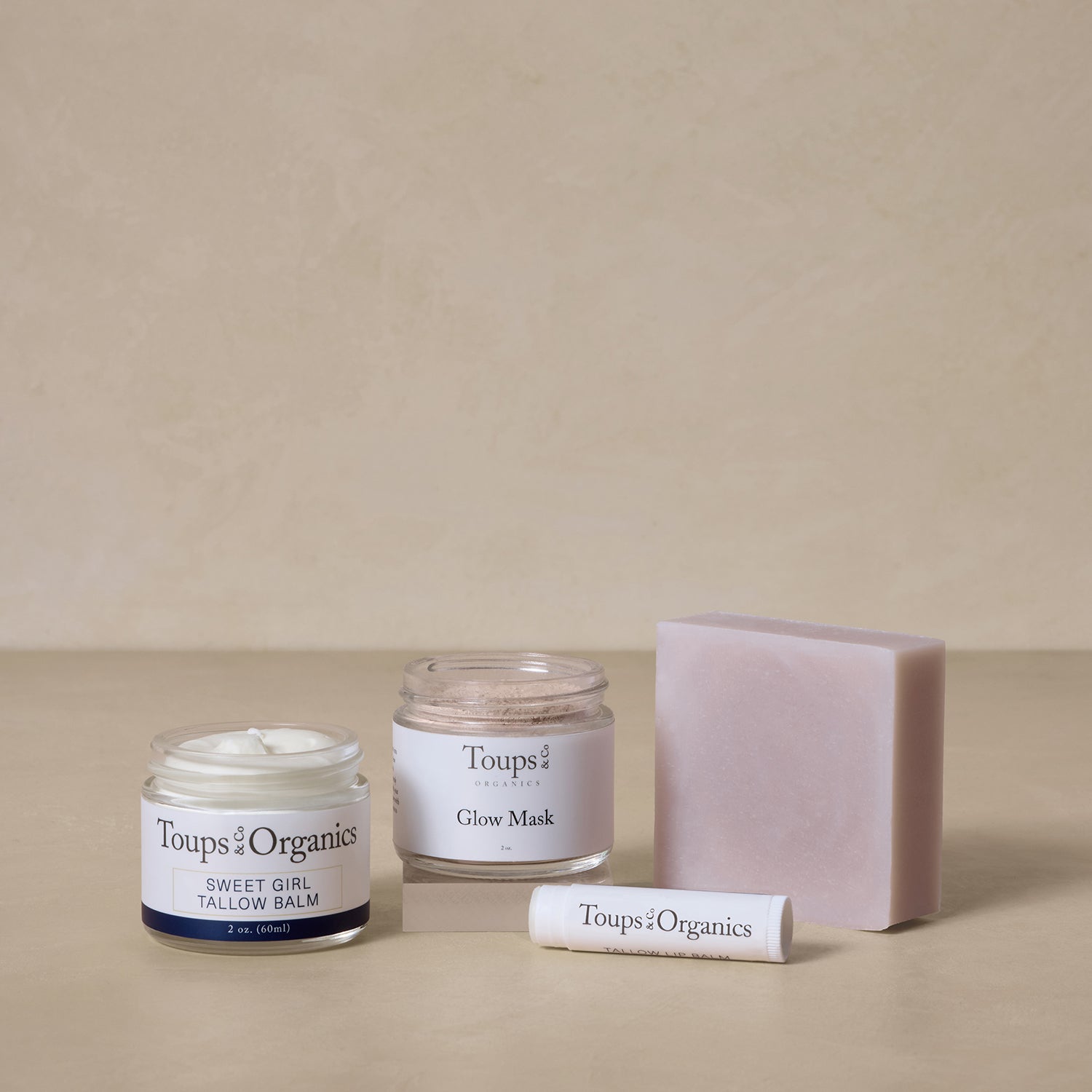 Four Toups and Co Organics Spa Gift Set skincare products—a tallow balm, glow mask, bar soap, and lip balm—rest on a beige surface; each item is nontoxic and perfect for skin hydration or as a thoughtful gift.