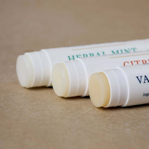 Three uncapped Tallow Lip Love tubes from Toups and Co Organics sit on a tan surface, each featuring distinctively colored labels highlighting their ultra-hydrating flavors.