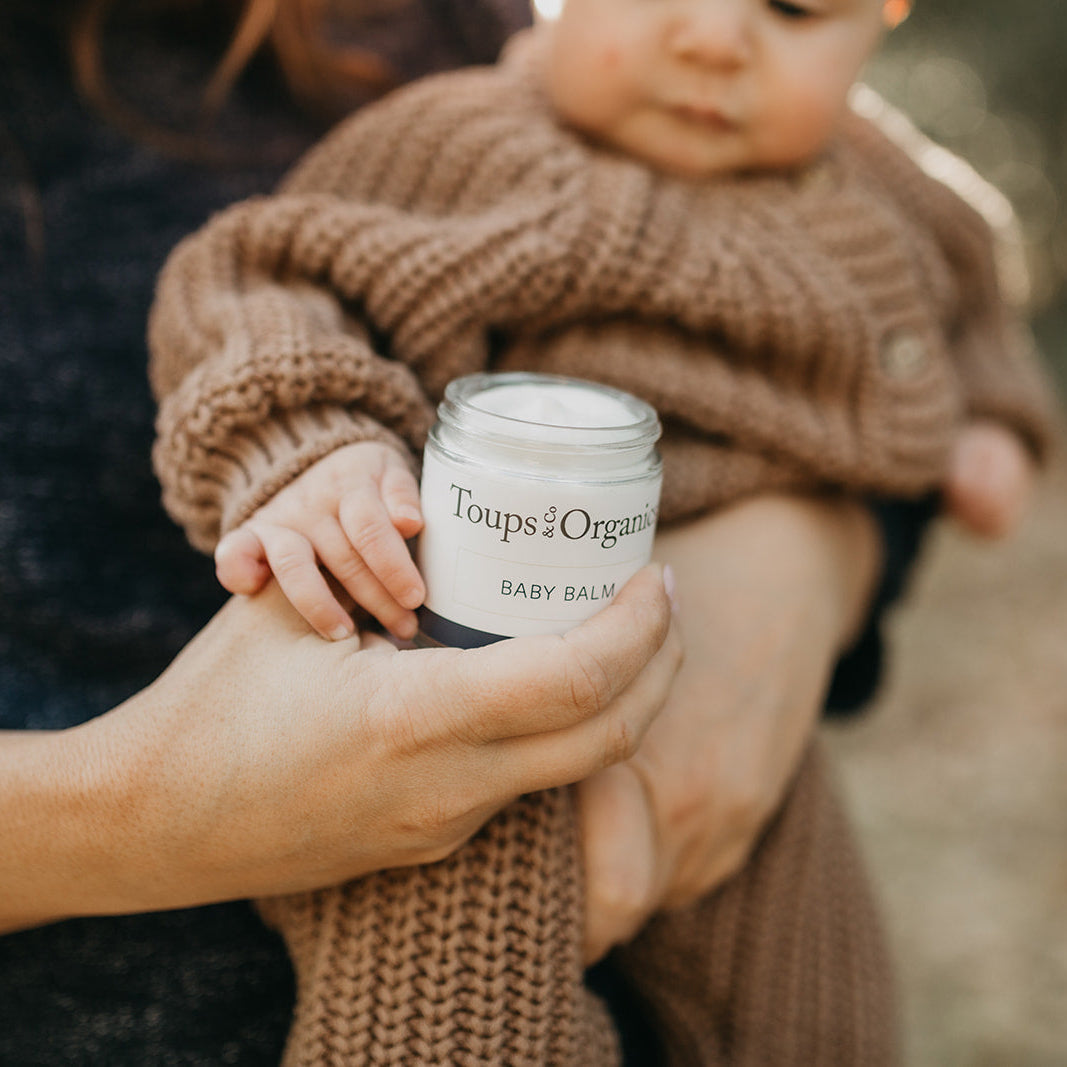 A parent holds Toups and Co Organics Baby Balm while cuddling a baby in a brown knit outfit—the ideal balm for easing eczema or diaper rash.