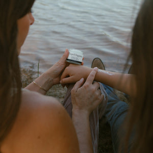Two people sit by the water; one applies Toups and Co Organics Tallow Balm - Sweet Girl from a small jar to the other’s arm, providing deep moisture and skin nourishment.