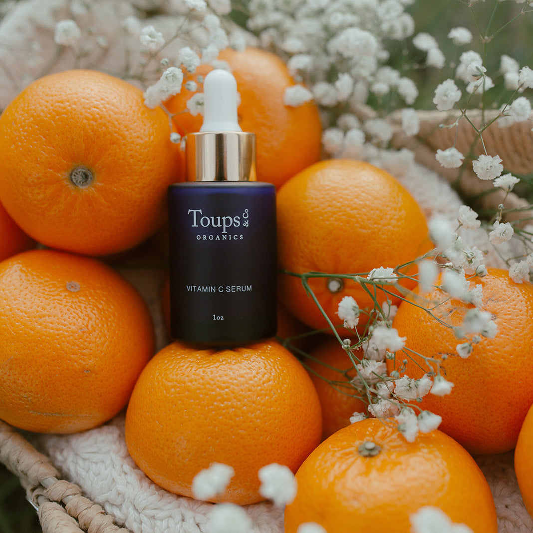 A bottle of Toups and Co Organics Vitamin C Serum sits among fresh oranges and white flowers in a woven basket, emphasizing its antioxidant benefits for brightening dull skin.