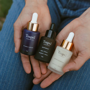 A person holds three Toups Organics serum bottles—Vitamin C Serum, Glow Serum, and Repairative Serum—in their hands, with blue jeans visible in the background.