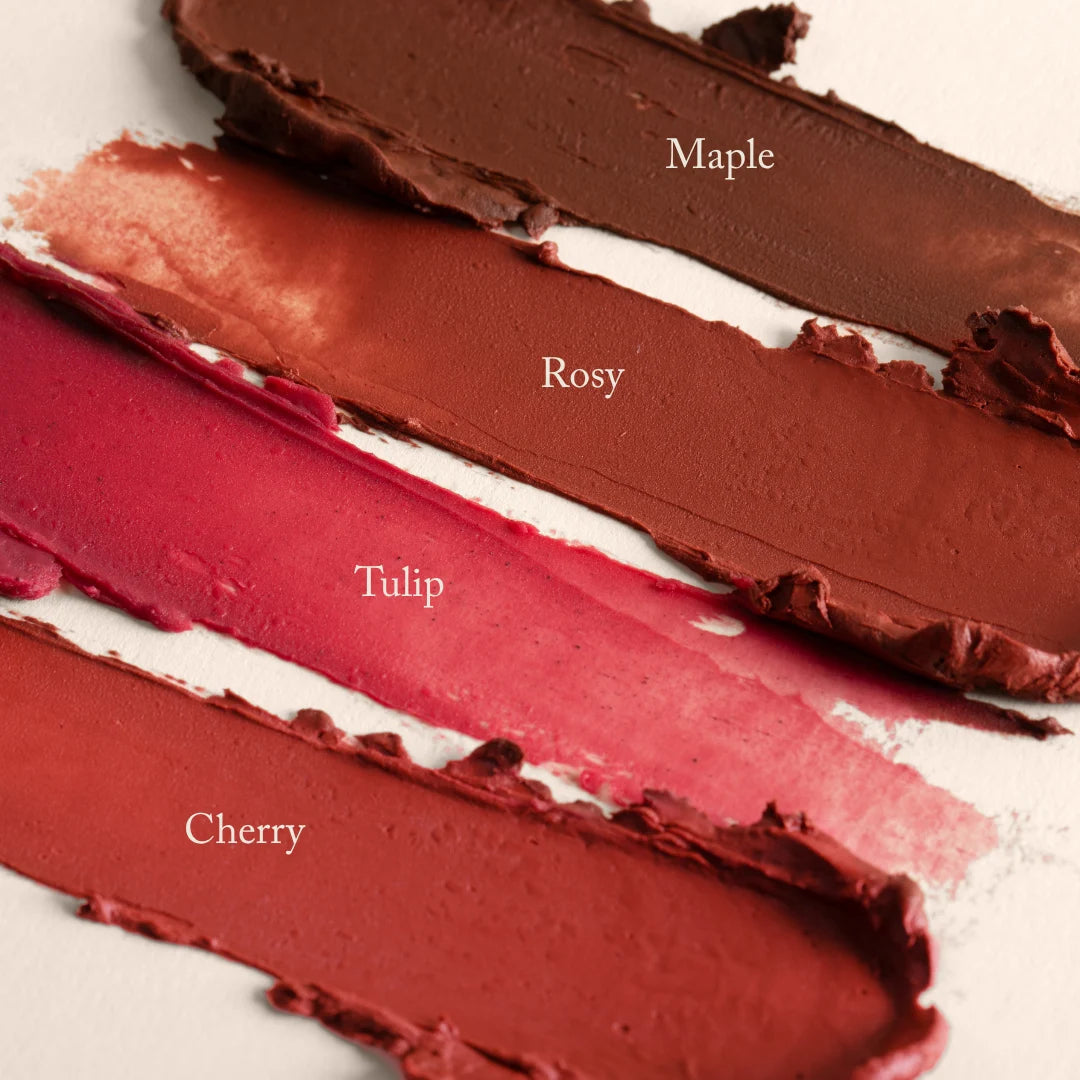 Four swatches from the Toups and Co Organics Complete Makeup Set—Maple, Rosy, Tulip, and Cherry—display a range of red and brown shades on a beige surface.