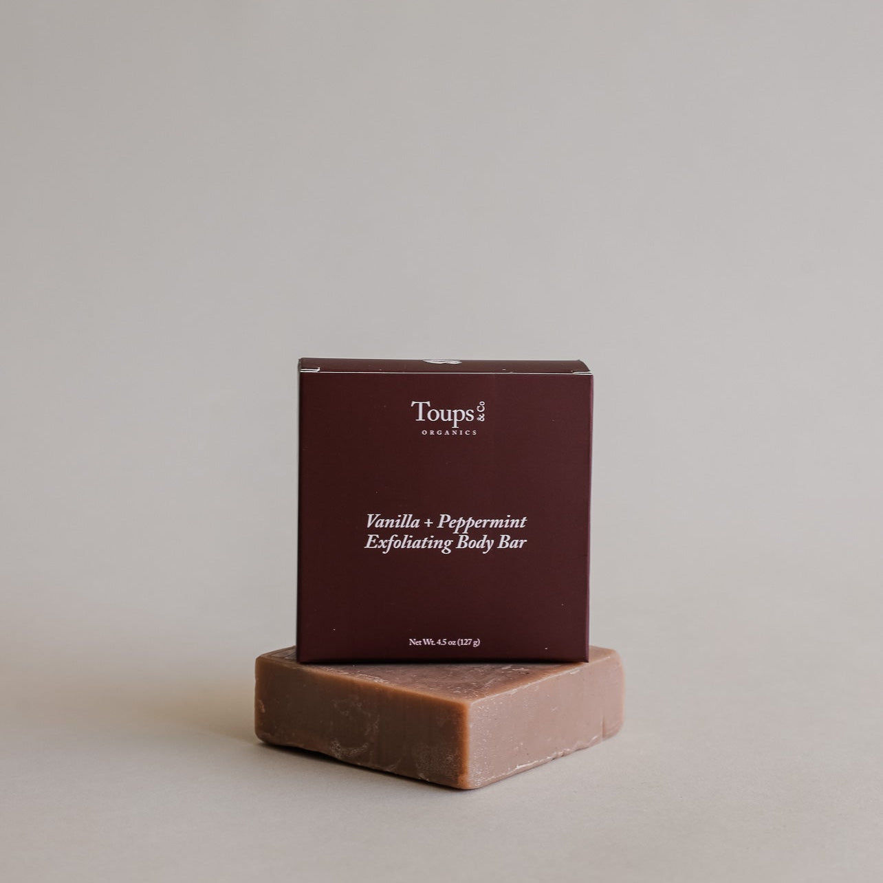 A box labeled Vanilla Peppermint Exfoliating Body Bar by Toups and Co Organics stands atop an unwrapped brown soap bar.