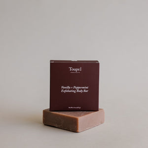 A box labeled Vanilla Peppermint Exfoliating Body Bar by Toups and Co Organics stands atop an unwrapped brown soap bar.