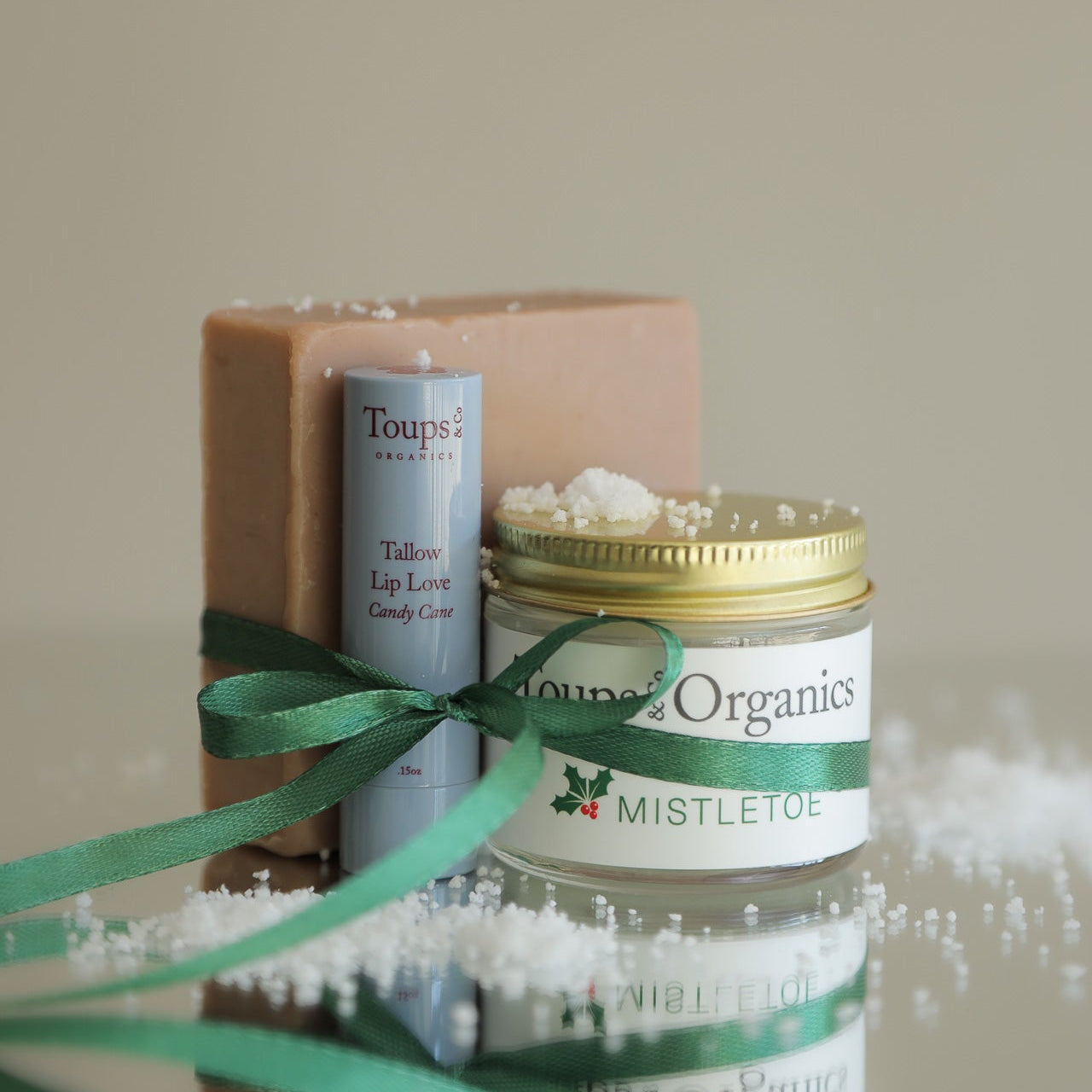 The Merry and Bright Gift Set by Toups and Co Organics features a bar of soap, lip balm, and a “Mistletoe” jar, all festively packaged with a green ribbon—an ideal holiday treat for natural skincare enthusiasts.