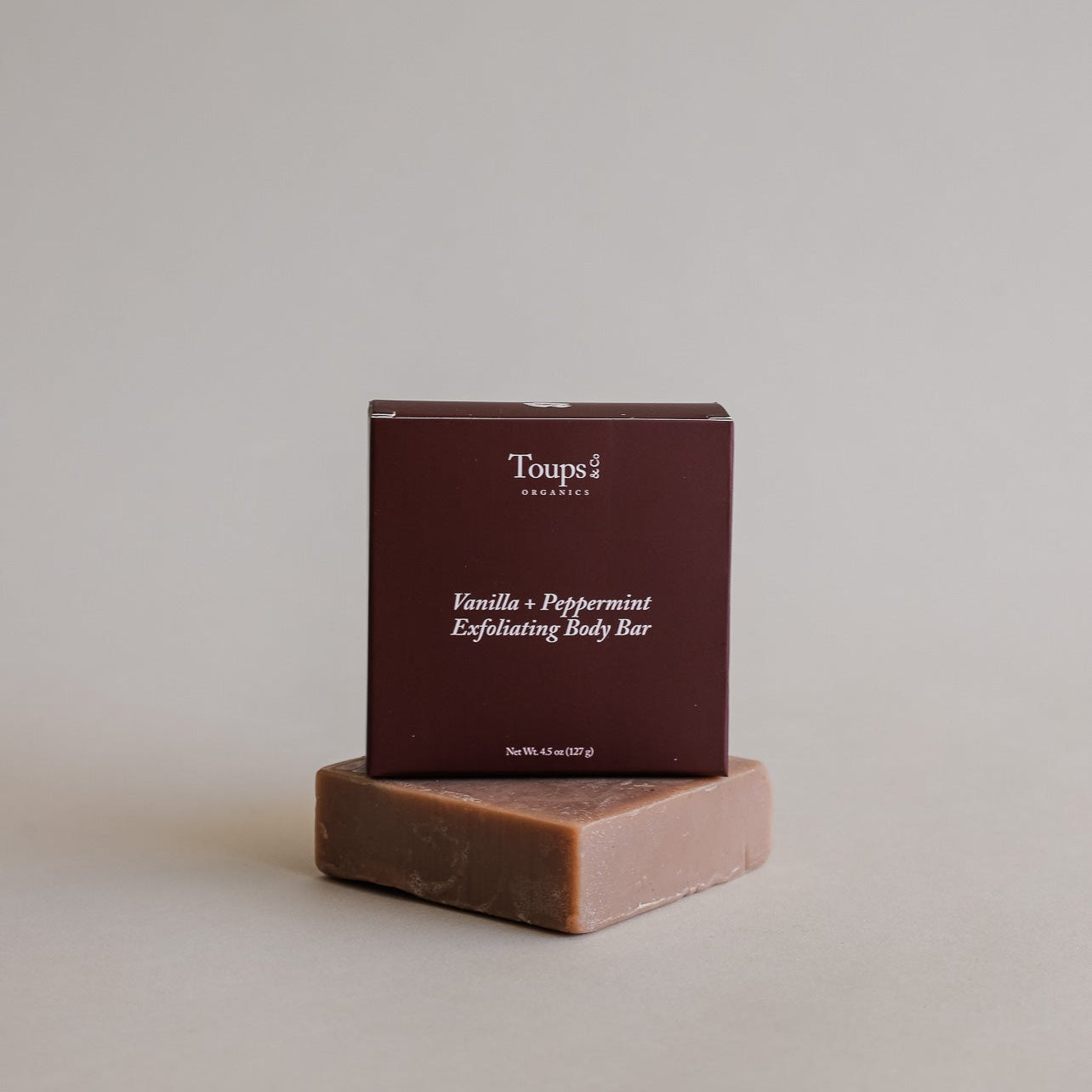 A brown bar of soap rests before a matching box labeled Toups and Co Organics Vanilla Peppermint Exfoliating Body Bar on a neutral background.