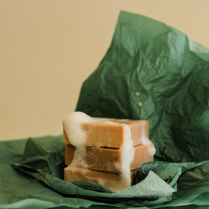 Two Toups and Co Organics Vanilla Peppermint Exfoliating Body Bars, topped with foam, are stacked on green crumpled tissue paper against a plain background.