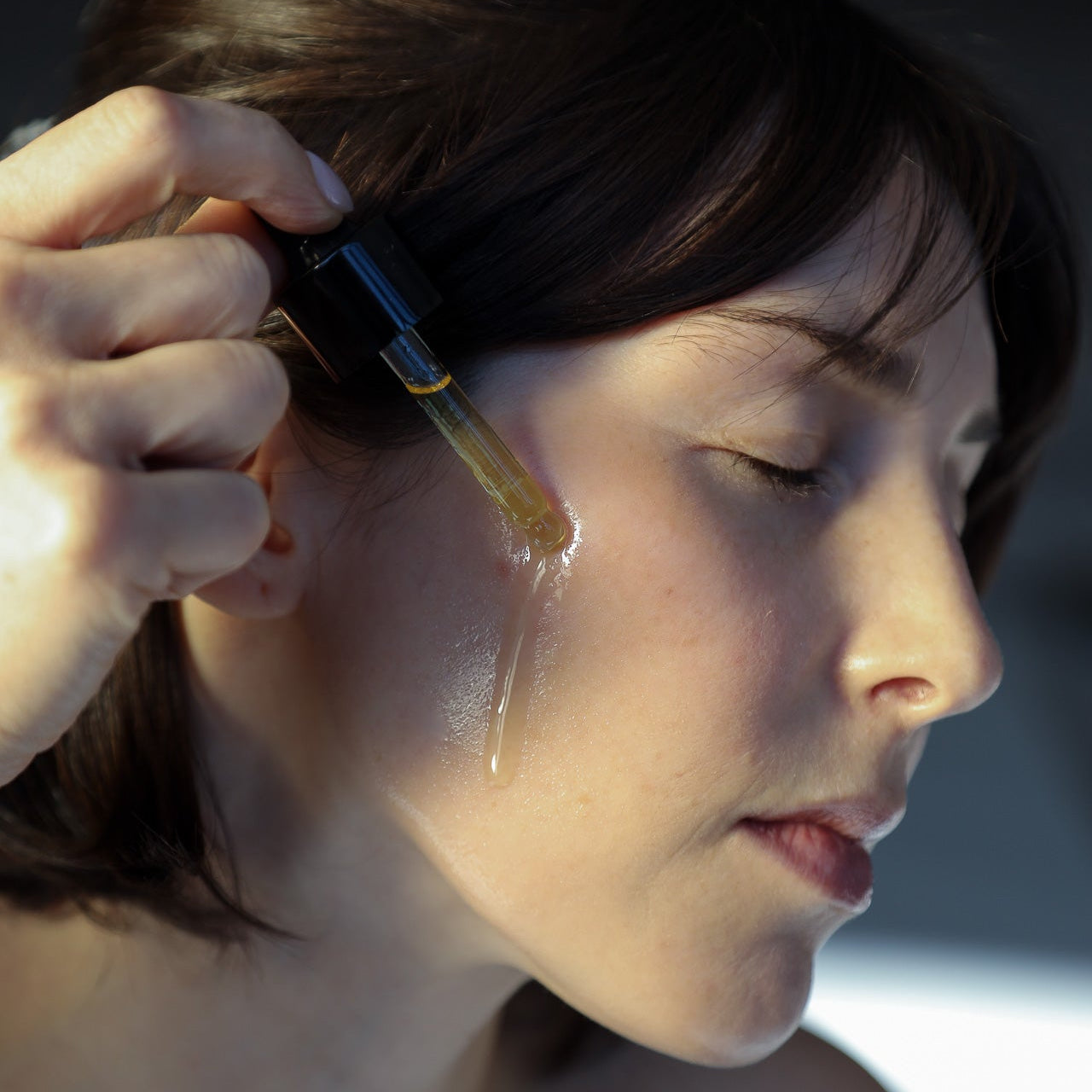 A person uses the Toups and Co Organics Glow Serum, applying it from a dropper onto their cheek with eyes closed to nourish their skin and enhance a healthy, radiant complexion.