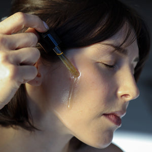 A person uses the Toups and Co Organics Glow Serum, applying it from a dropper onto their cheek with eyes closed to nourish their skin and enhance a healthy, radiant complexion.