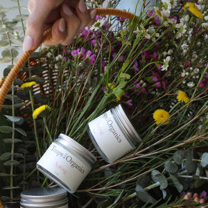 A hand holds a wicker basket of wildflowers, magnolia flowers, and three jars of Toups and Co Organics Spring Balm - Floral + Citrus.