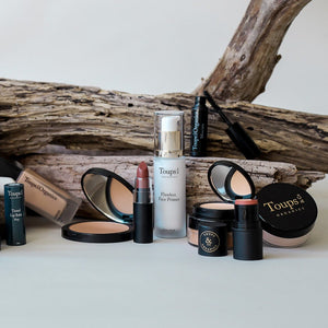 A selection of Toups and Co Organics makeup products—including the Flawless Face Primer, which is suitable for all skin types—along with lipstick, powders, and mascara, are displayed in front of driftwood pieces on a white surface.