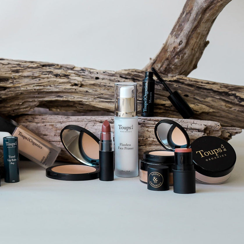 A selection of Toups and Co Organics makeup products—including the Flawless Face Primer, which is suitable for all skin types—along with lipstick, powders, and mascara, are displayed in front of driftwood pieces on a white surface.