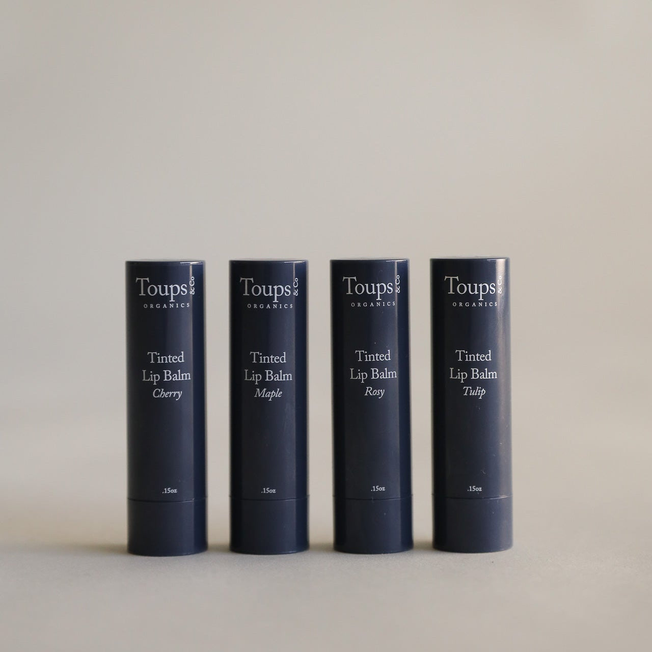 Four dark blue tubes from the Toups and Co Organics Complete Makeup Set, labeled Cherry, Maple, Rosy, and Tulip, stand upright in a row against a plain neutral background.