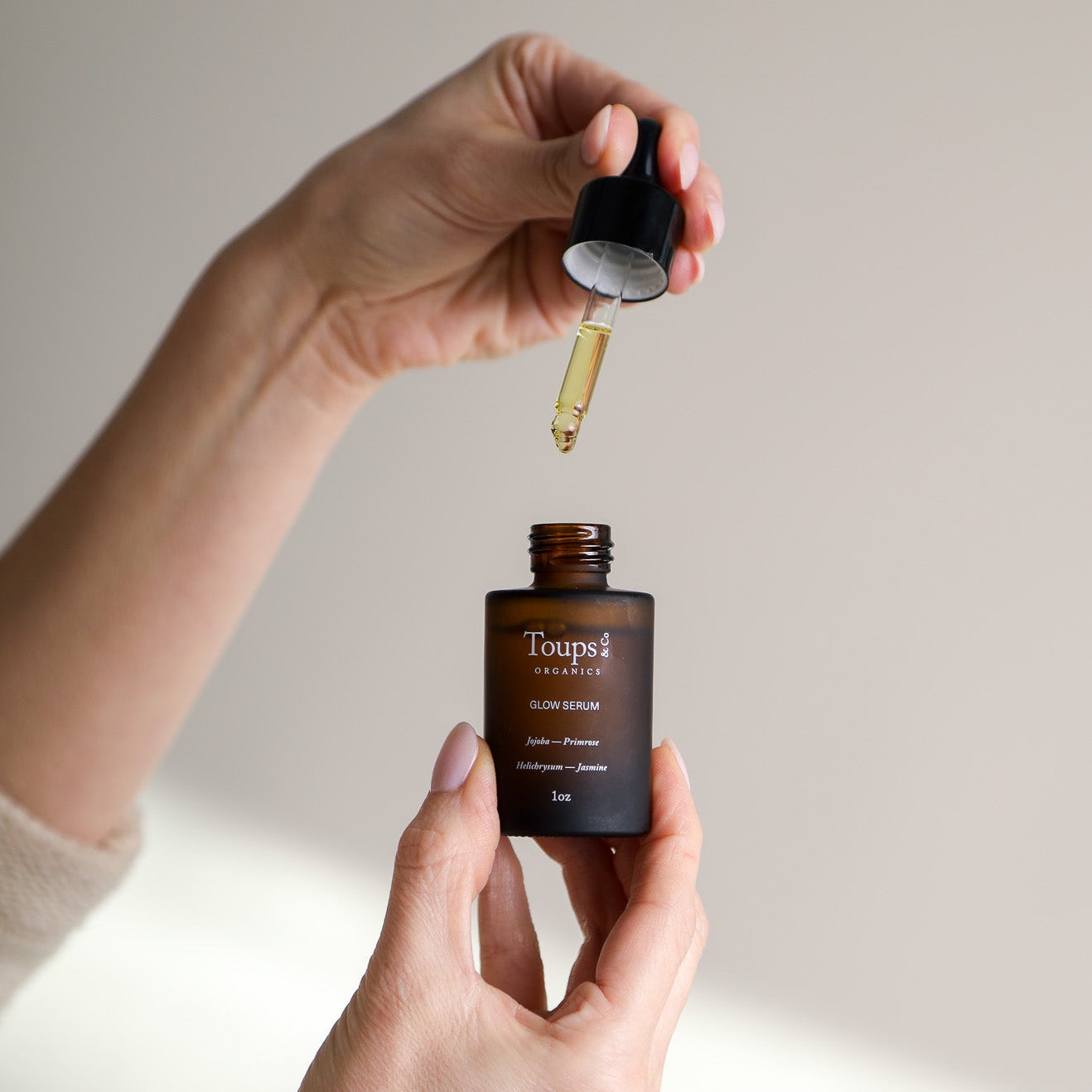 A person holds a dropper above an open bottle labeled Toups and Co Organics Glow Serum, displaying this healing serum against a neutral background.